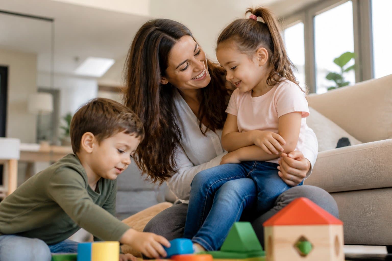 Mulher sorrindo com dois filhos pequenos em uma sala de estar iluminada e aconchegante.